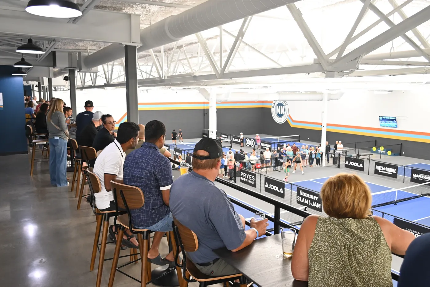 Guests on walnut stools at the mezzanine drink rail, a pint on the table, watching a tournament match and the crowd along the court fence below the Pints & Paddle MN logo wall