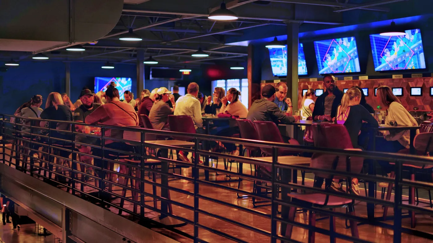 The mezzanine packed on a weeknight: high-top tables under blue and magenta lighting, a row of TVs and the lit tap wall along the right