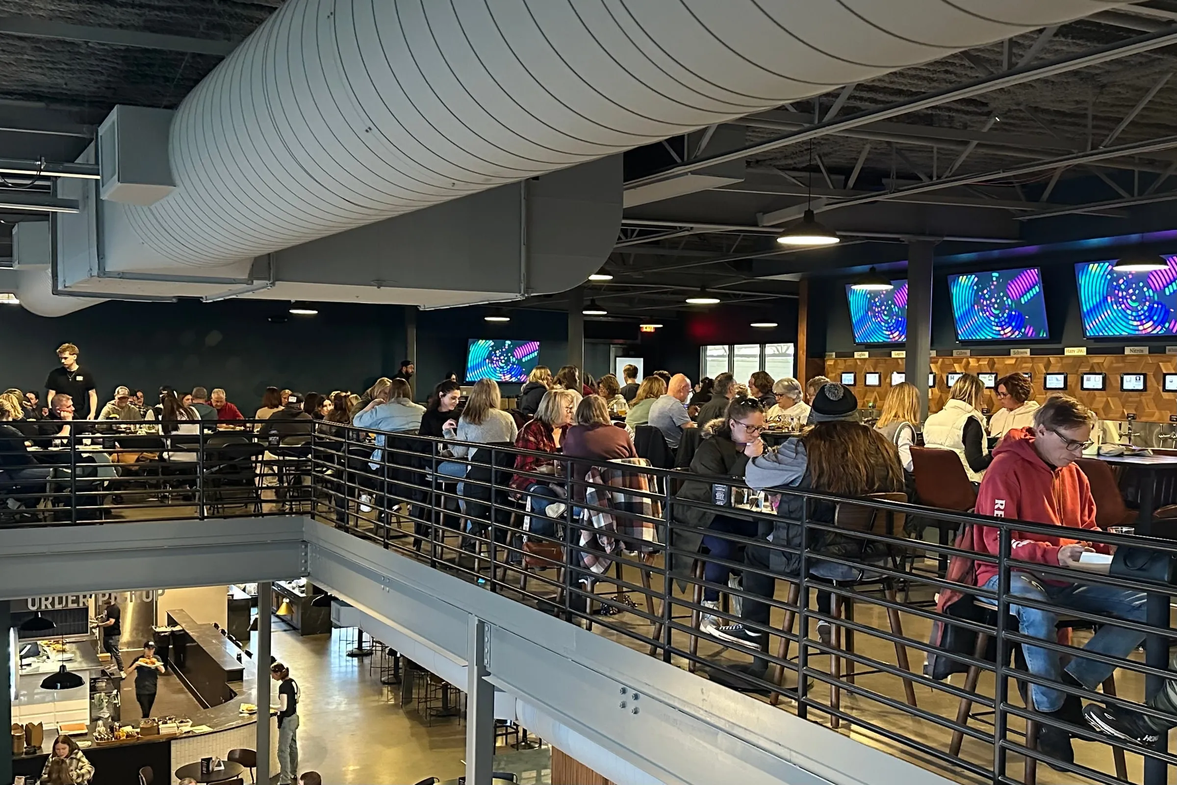 A packed mezzanine on a game night: tables of players along the rail, three screens lit over the upstairs tap wall, the order counter visible below