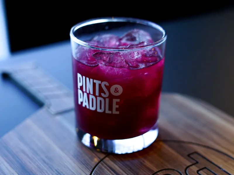A deep purple blueberry mint mocktail over ice in a Pints & Paddle rocks glass resting on a wooden pickleball paddle