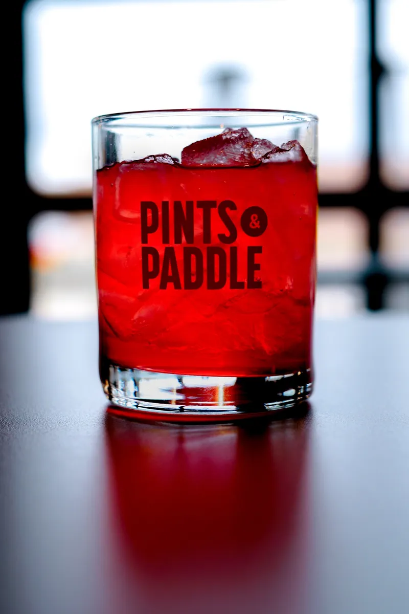 A bright red cranberry ginger mocktail over ice in a Pints & Paddle logo rocks glass in front of a bright window
