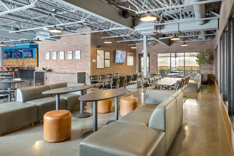 North mezzanine lounge from the window side: grey sofas and cognac ottomans around oval tables, long tables with black leather chairs, framed prints and a TV on the brick wall