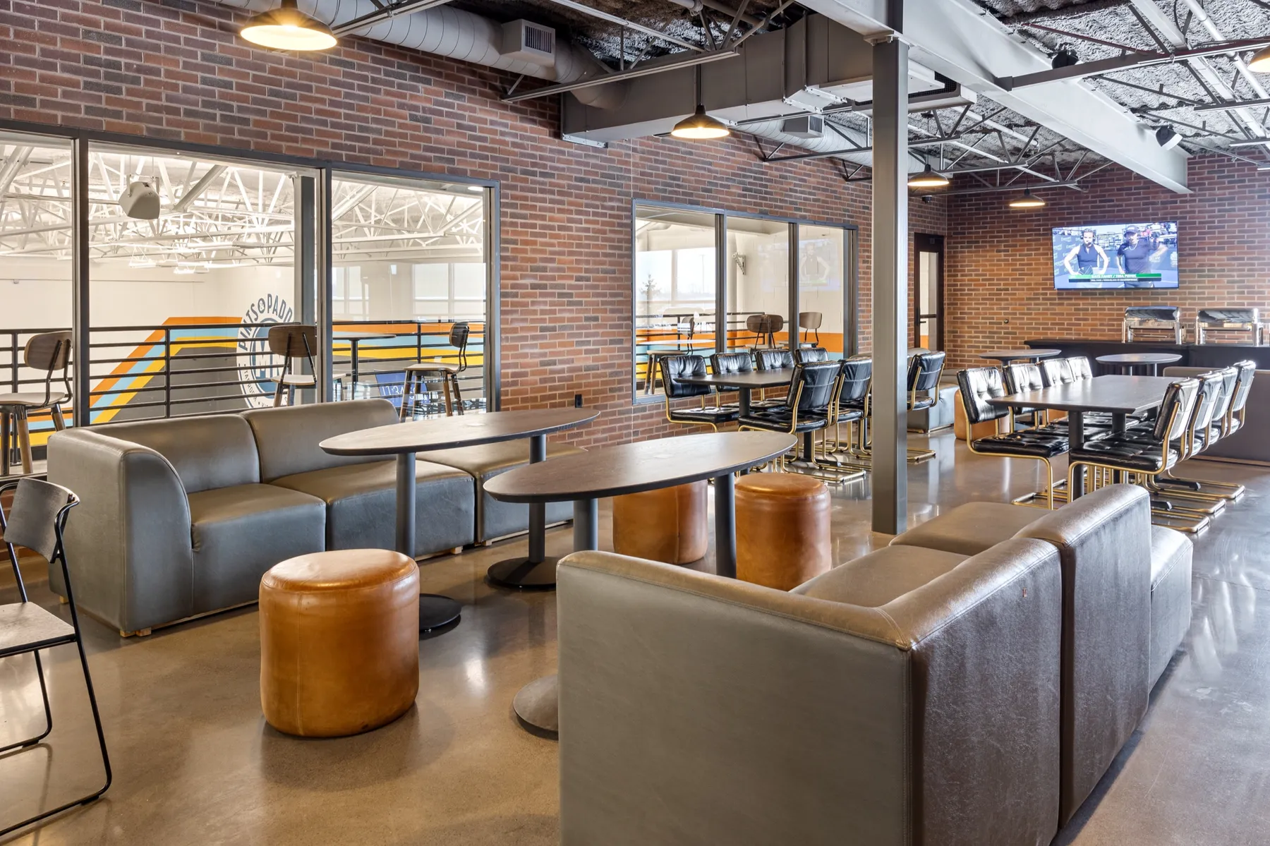 North mezzanine lounge with grey sofas and cognac ottomans around oval tables, long dining-height tables with black leather chairs, and a wall TV over the buffet counter