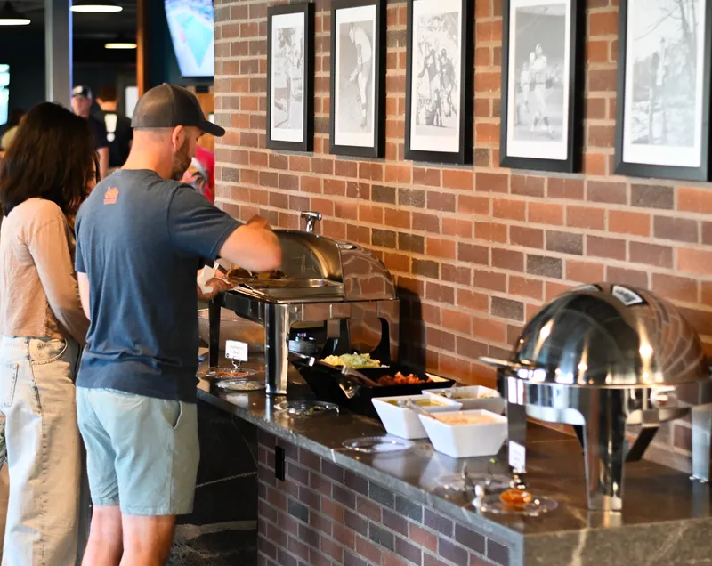 Guests serving themselves barbacoa from a chafing dish on the north mezzanine granite buffet counter, under the framed black-and-white prints on the brick wall, with the high-tops of the neighboring mezzanine seating and the tap-wall TVs behind