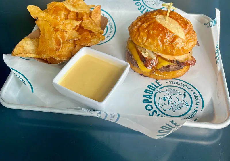 The beer cheese burger on a pretzel roll with a cup of beer cheese and kettle chips on a Pints & Paddle tray
