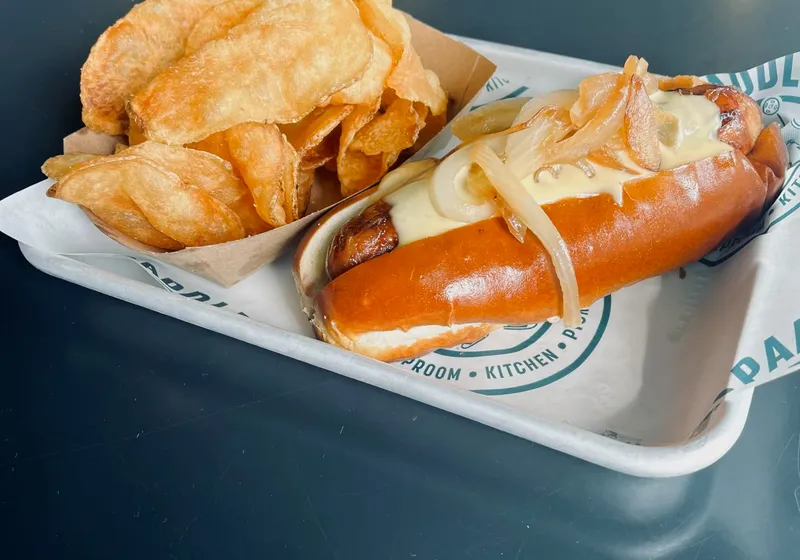 A bratwurst on a pretzel roll topped with caramelized onion and cheese, with kettle chips, on Pints & Paddle paper