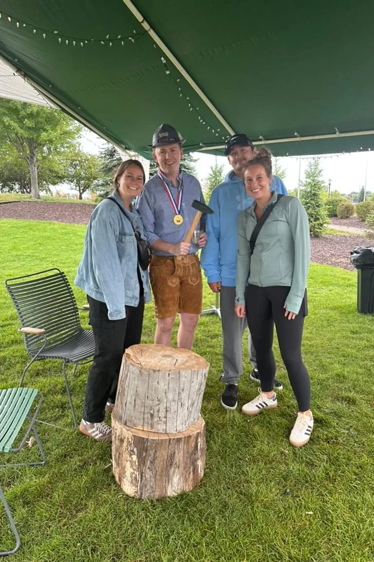 Three guests around the Hammerschlagen stump on the lawn, one in lederhosen holding the hammer