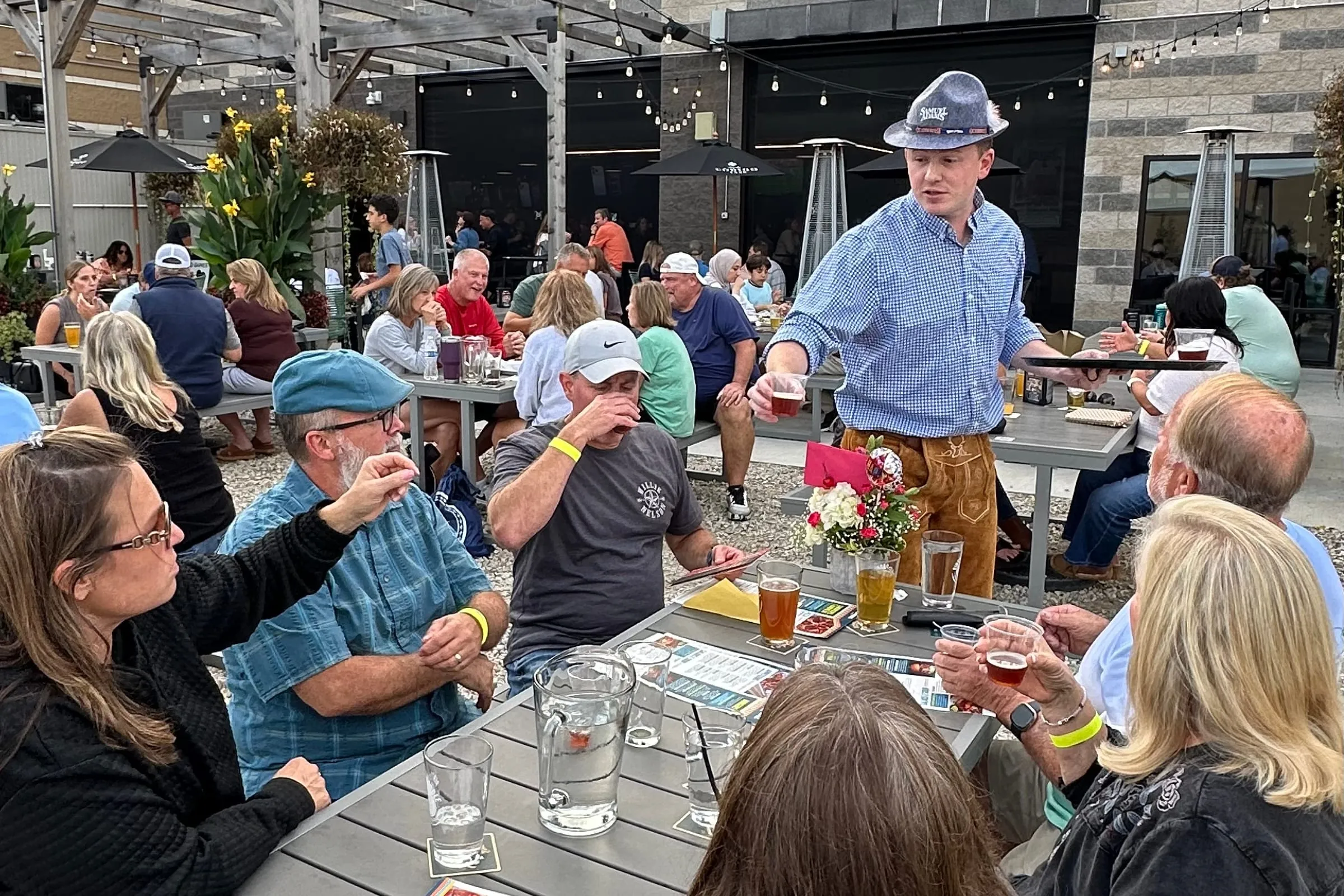 A full patio at Oktoberfest, a host in a Bavarian hat and lederhosen pouring at a table of guests with beers