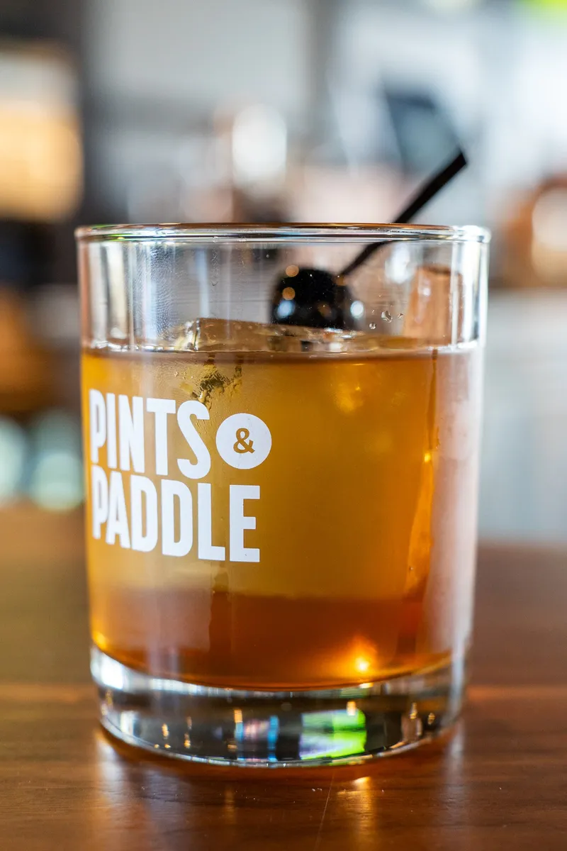 An amber Orchard Old Fashioned over a large ice cube in a Pints & Paddle rocks glass, with a dark cherry on a pick.