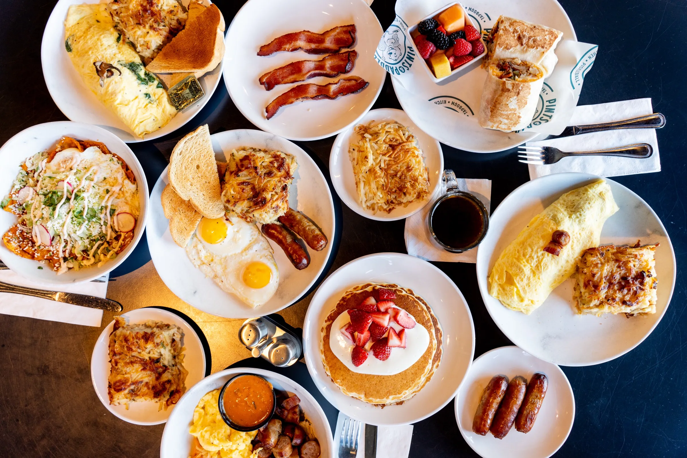 The full weekend breakfast spread from above: omelettes, bacon, a breakfast wrap with fruit, chilaquiles, pancakes with strawberries and a cup of coffee