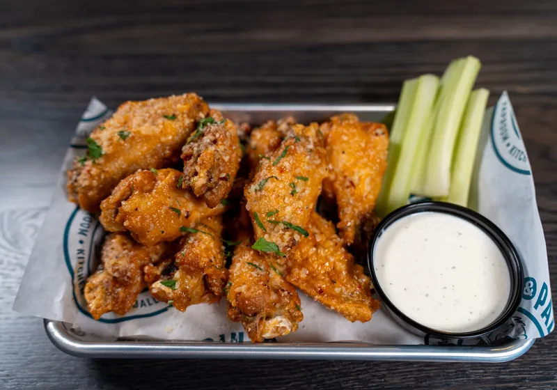 A metal tray of parmesan garlic bone-in wings dusted with parsley, with celery sticks and a ramekin of ranch