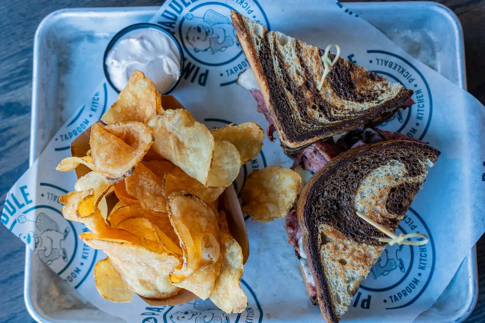 Two halves of a pastrami sandwich on toasted marble rye beside kettle chips and a cup of sauce.