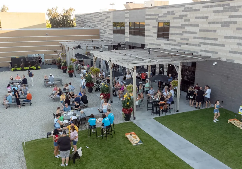 Elevated view of the patio at golden hour, with guests under the pergola, picnic tables on the gravel and cornhole on the lawn