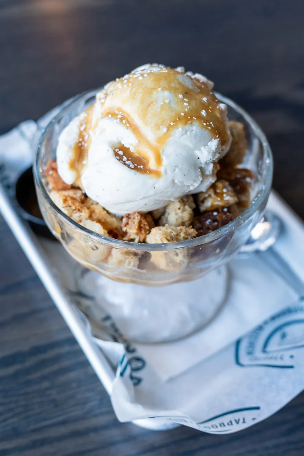 Peach cobbler in a glass dessert dish topped with vanilla ice cream, caramel and a dusting of powdered sugar.