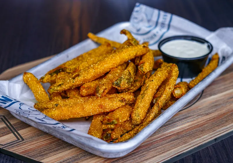 Cornmeal-breaded pickle fries piled in a metal tray with a cup of ranch, on branded paper over a wooden board