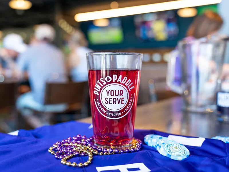 A Your Serve pint glass of red beer and a full pitcher on a purple jersey with beads, the bar behind