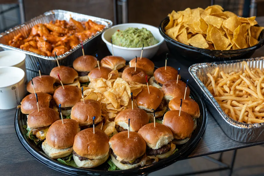 A catering spread of cheeseburger sliders, buffalo wings, fries, tortilla chips and guacamole in trays on a table