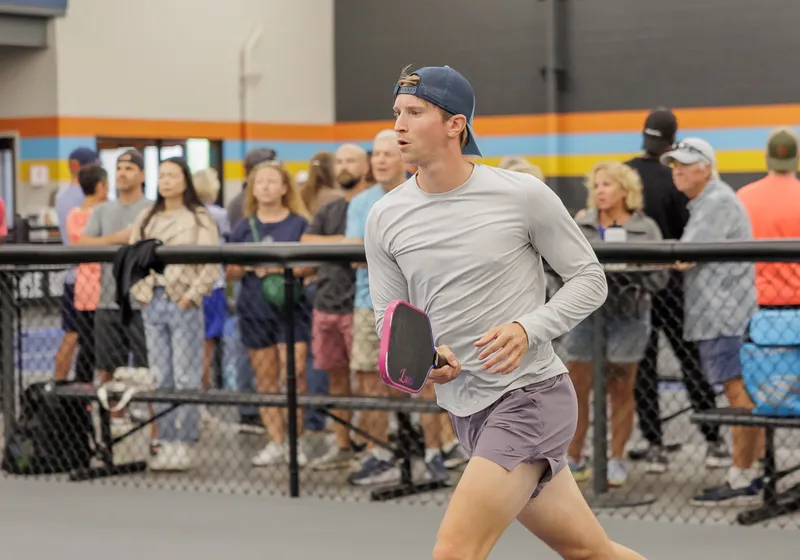 A player in a backwards cap sprints across the court with a pink paddle, spectators along the fence
