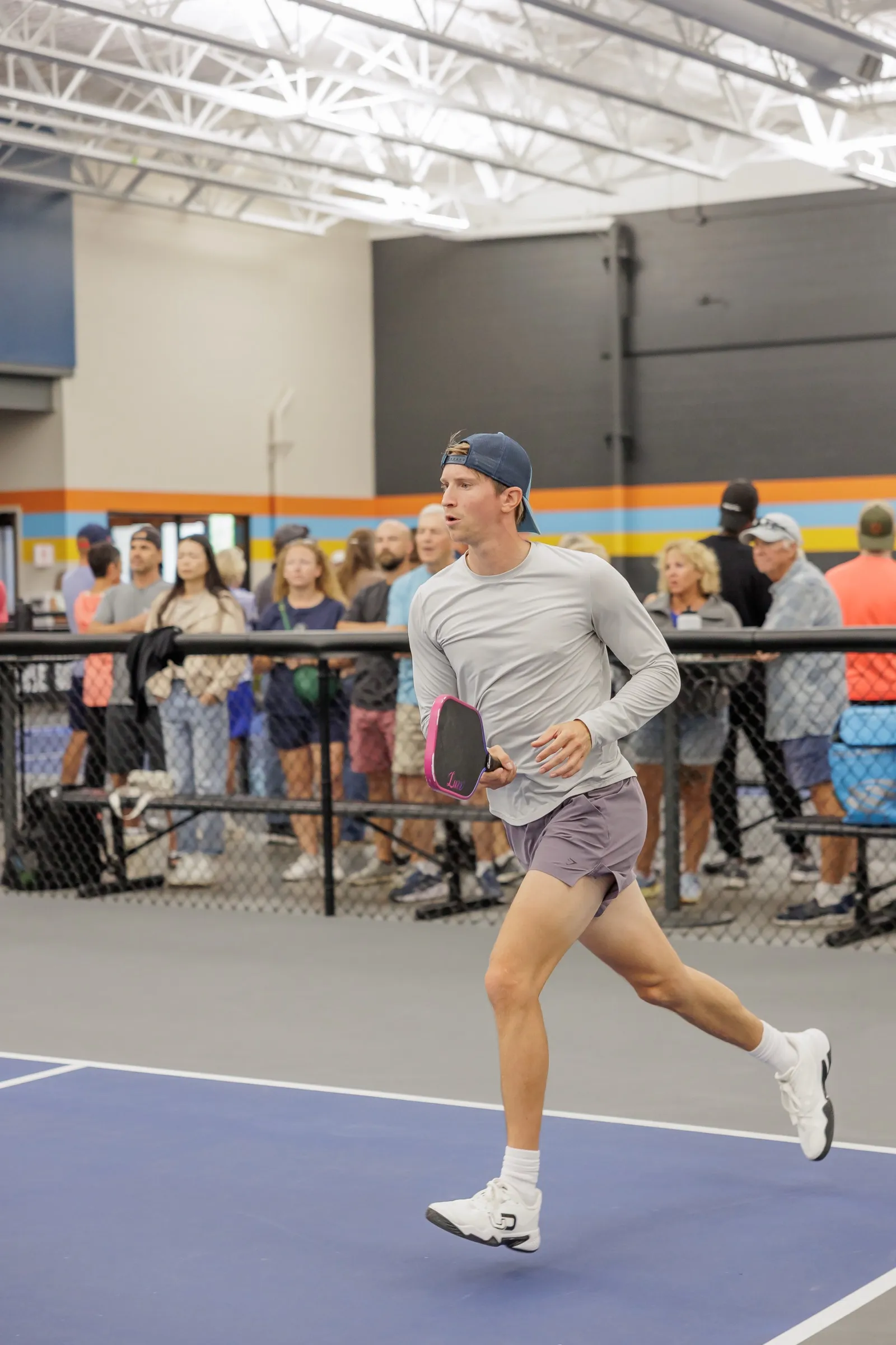 A player in a backwards cap sprinting across the court with a pink paddle, spectators lined along the fence behind him