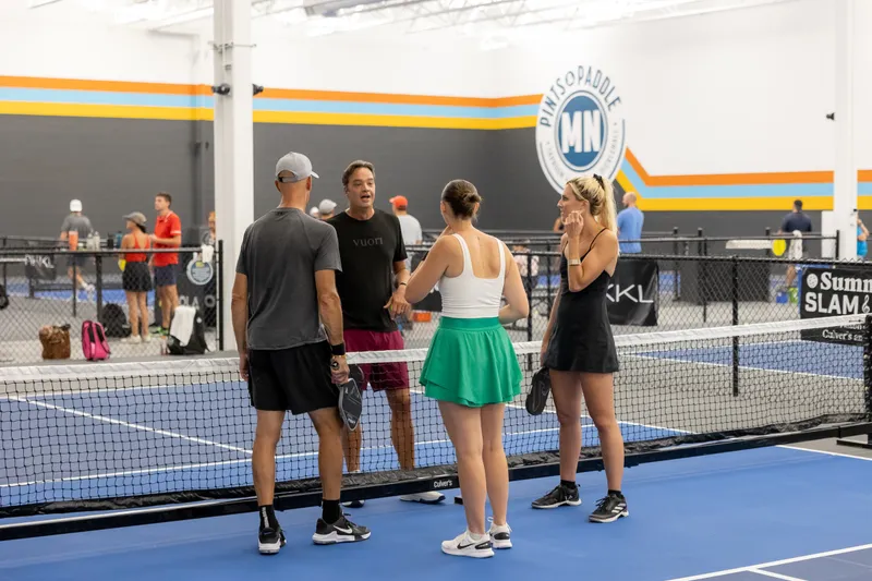 Four players chatting at the net on a blue indoor court under the Pints & Paddle logo wall