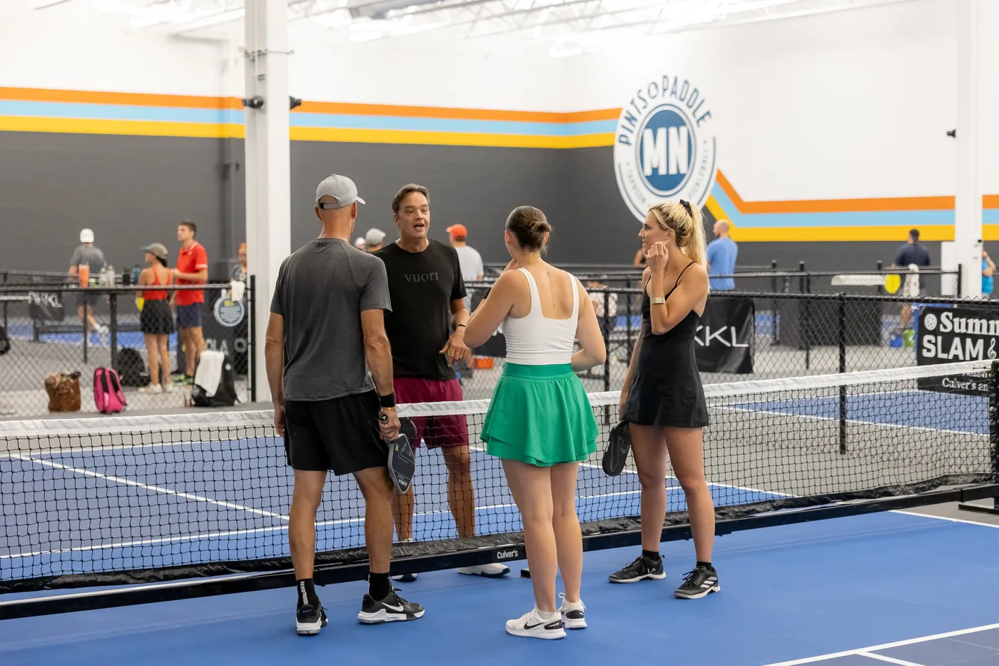 Four players chatting at the net on a blue indoor court beneath the Pints & Paddle MN logo wall