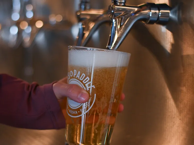 A hand in a plum sleeve holds a Pints & Paddle pint glass under a chrome self-serve tap as golden beer fills it to a foamy head