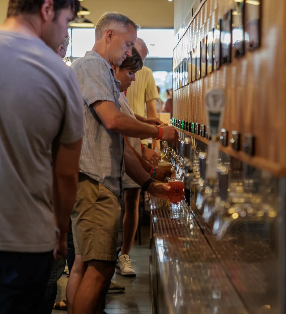 Guests lined up along the wooden self-serve tap wall, one pouring a beer under the rows of tap screens