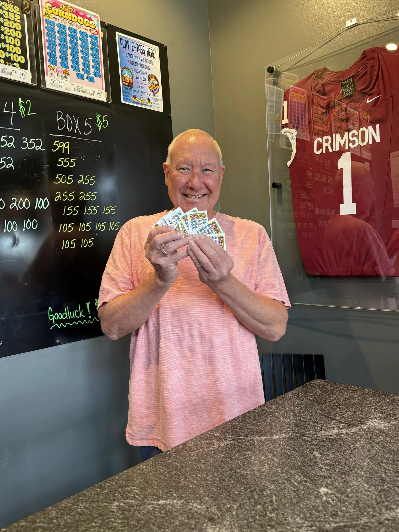 A smiling guest at the pull tab booth fanning out a handful of tickets, with the prize board, a Play E-Tabs Here sign and a framed Crimson jersey behind him
