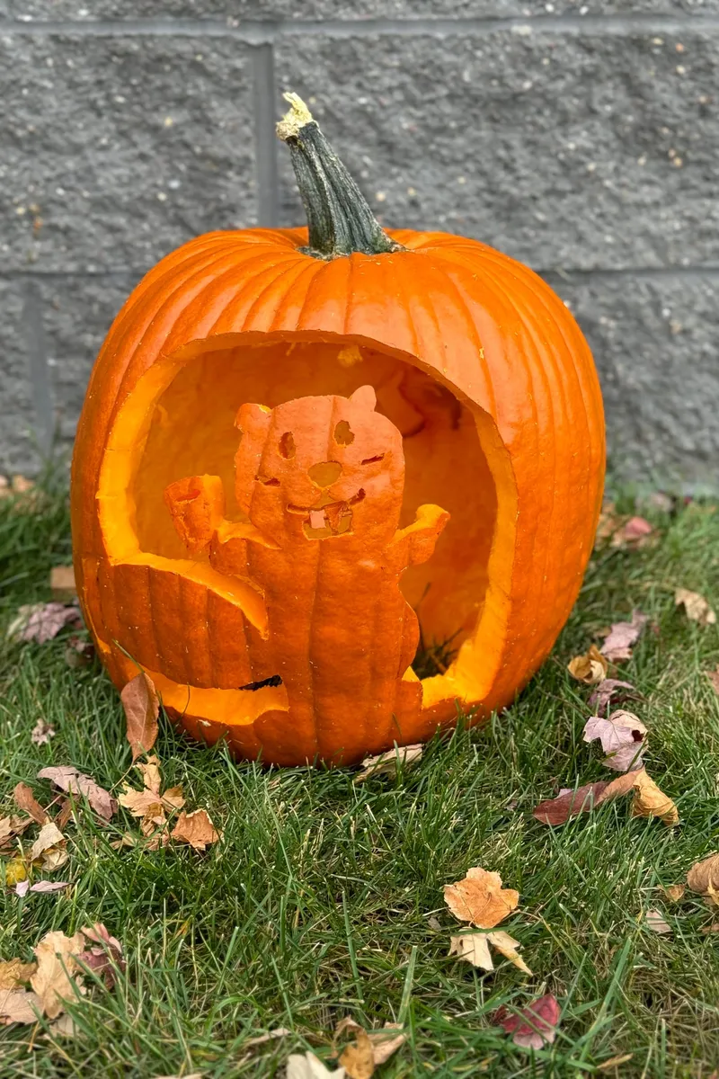 A pumpkin carved with a waving beaver, set on the grass among fallen leaves