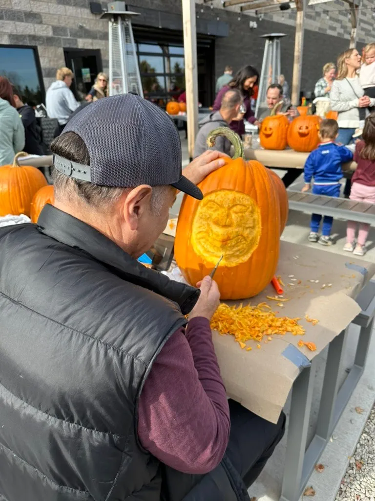 A man in a cap carving a detailed face into a tall pumpkin, shavings piled on the table