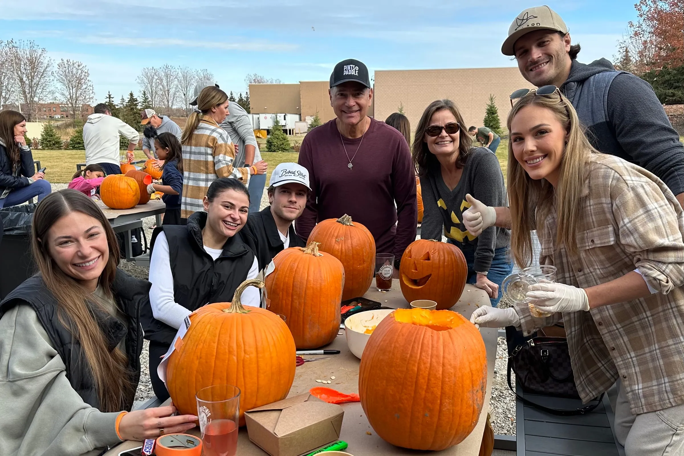 Six friends around a patio table with carved and uncarved pumpkins, one holding up a cider, gloves on