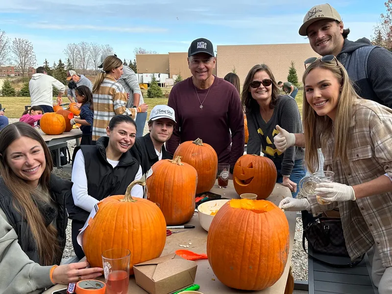 Six friends around a patio table with carved and uncarved pumpkins, one holding up a cider, gloves on