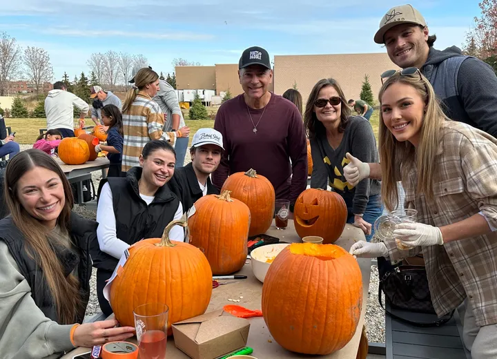 Six friends around a patio table with carved and uncarved pumpkins, one holding up a cider, gloves on