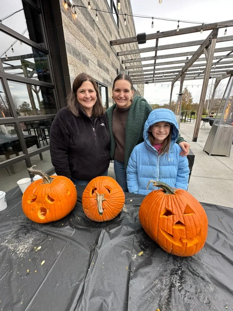 Two women and a girl in a blue coat behind their three finished jack-o'-lanterns on the patio