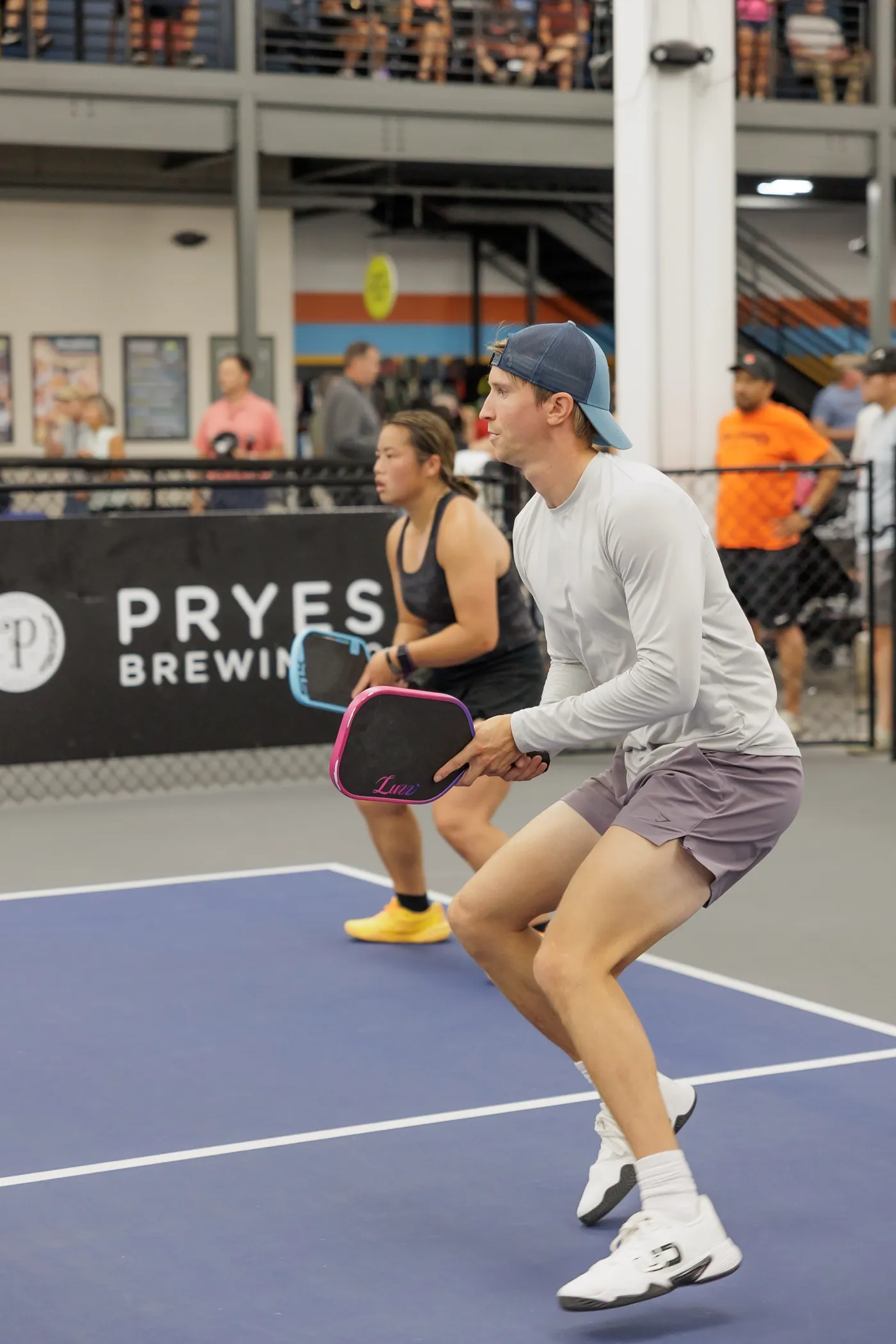 Two mixed-doubles players in a ready crouch on a blue indoor court with the mezzanine crowd above