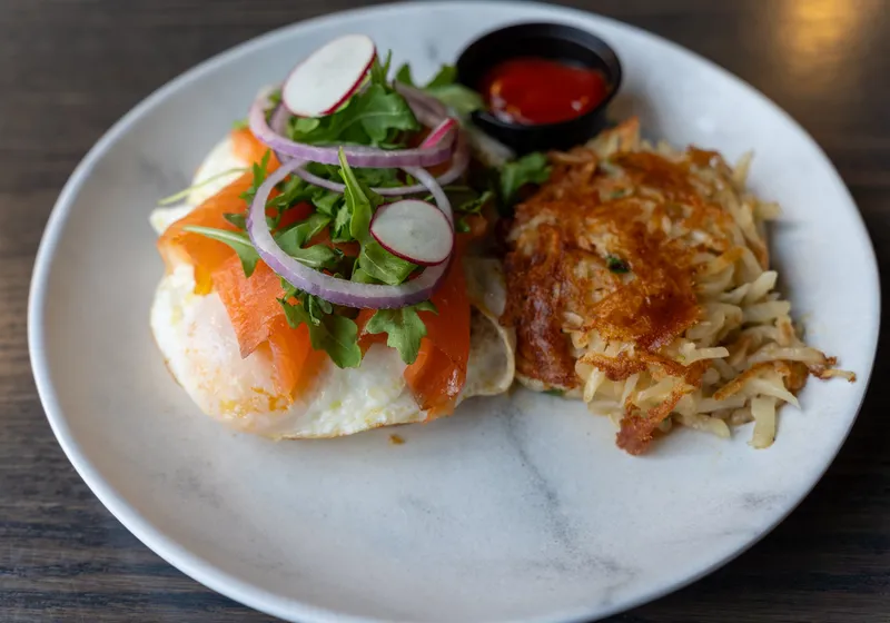 Smoked salmon toast topped with an egg, arugula, red onion and radish, beside a golden hash brown