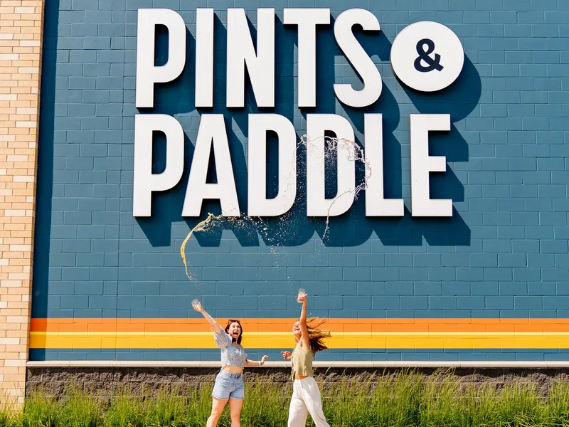 Two friends leap and toss their drinks in front of the Pints & Paddle sign