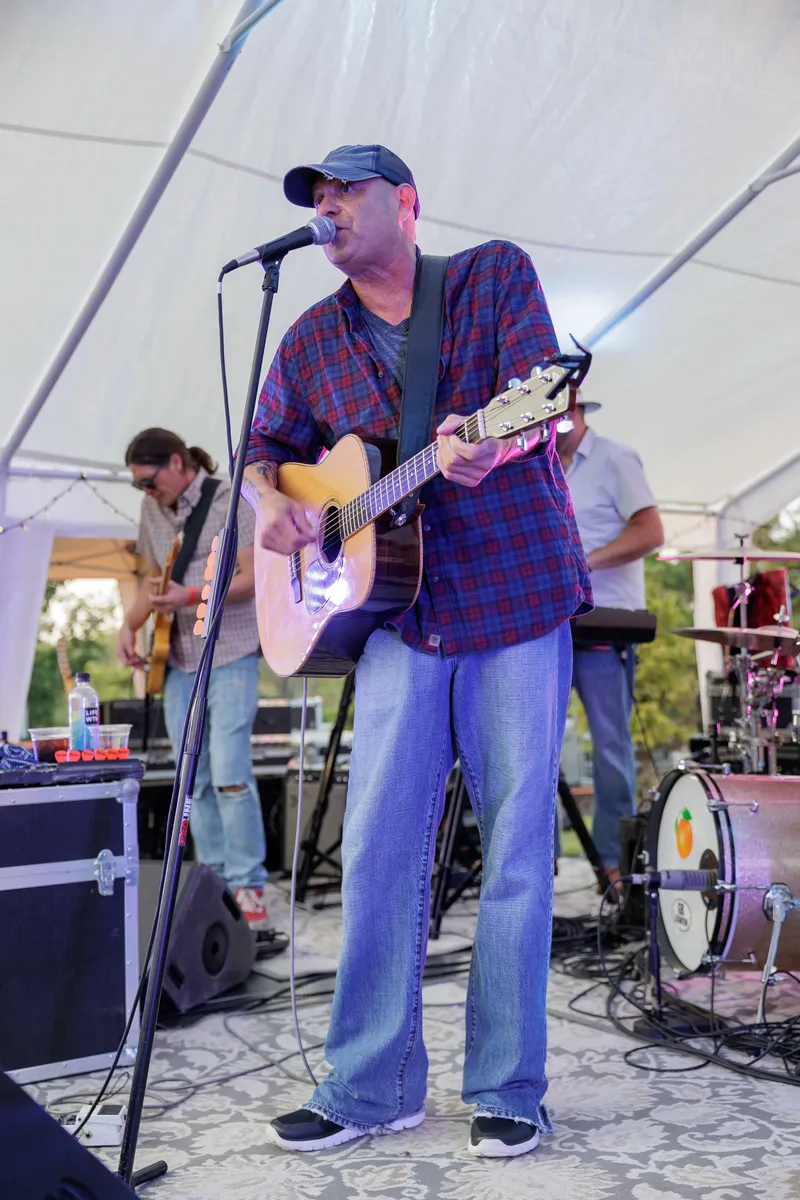 A singer-guitarist in a plaid shirt and cap at the mic under the white tent, keys and drums behind him