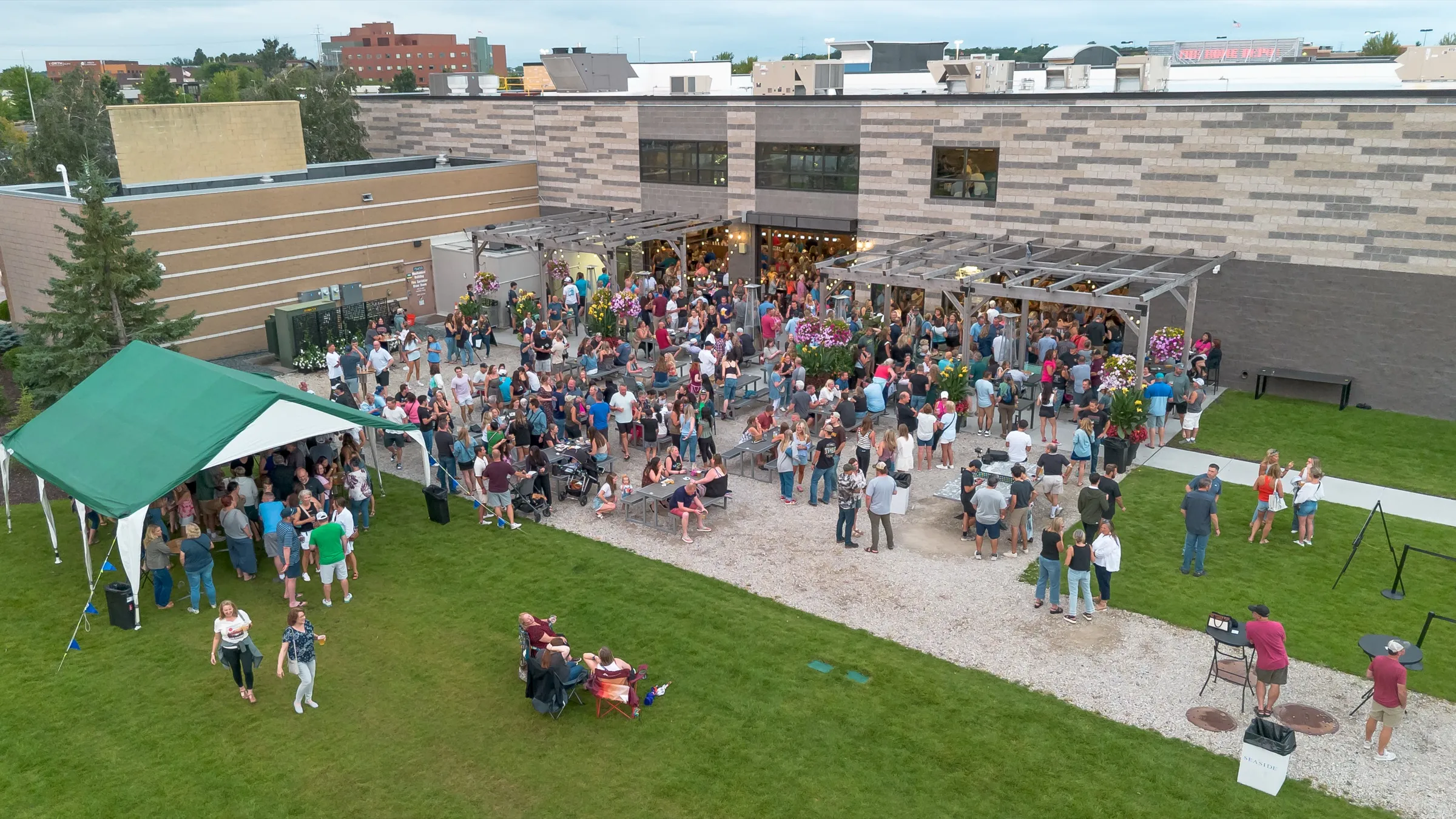 Wide aerial view of the patio and building during Slam & Jam, with a crowd under the pergolas and across the gravel and the Maple Grove skyline behind