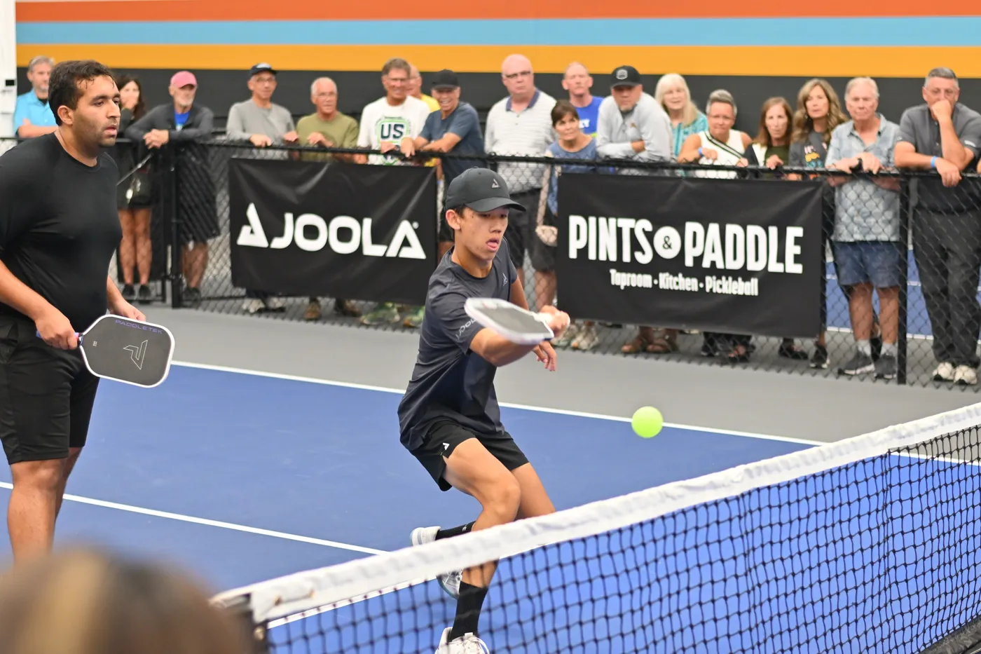 A pro lunges into a low forehand at the kitchen line, the ball just off his paddle, with spectators packed two deep along the fence behind the Pints & Paddle and JOOLA banners