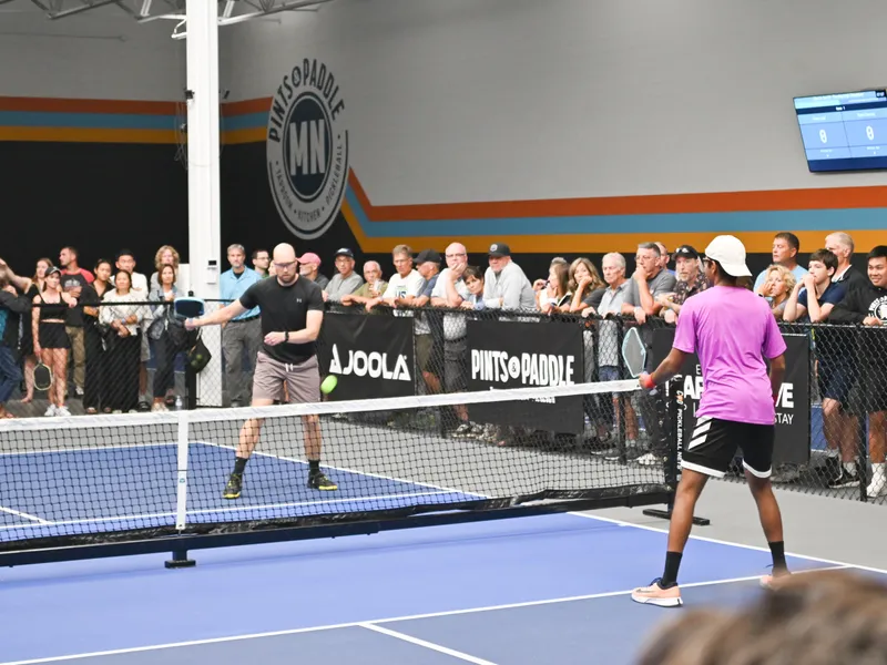 Two players square off at the net during Slam & Jam, the ball hanging over the tape and spectators two deep along the fence beneath the Pints & Paddle MN logo wall