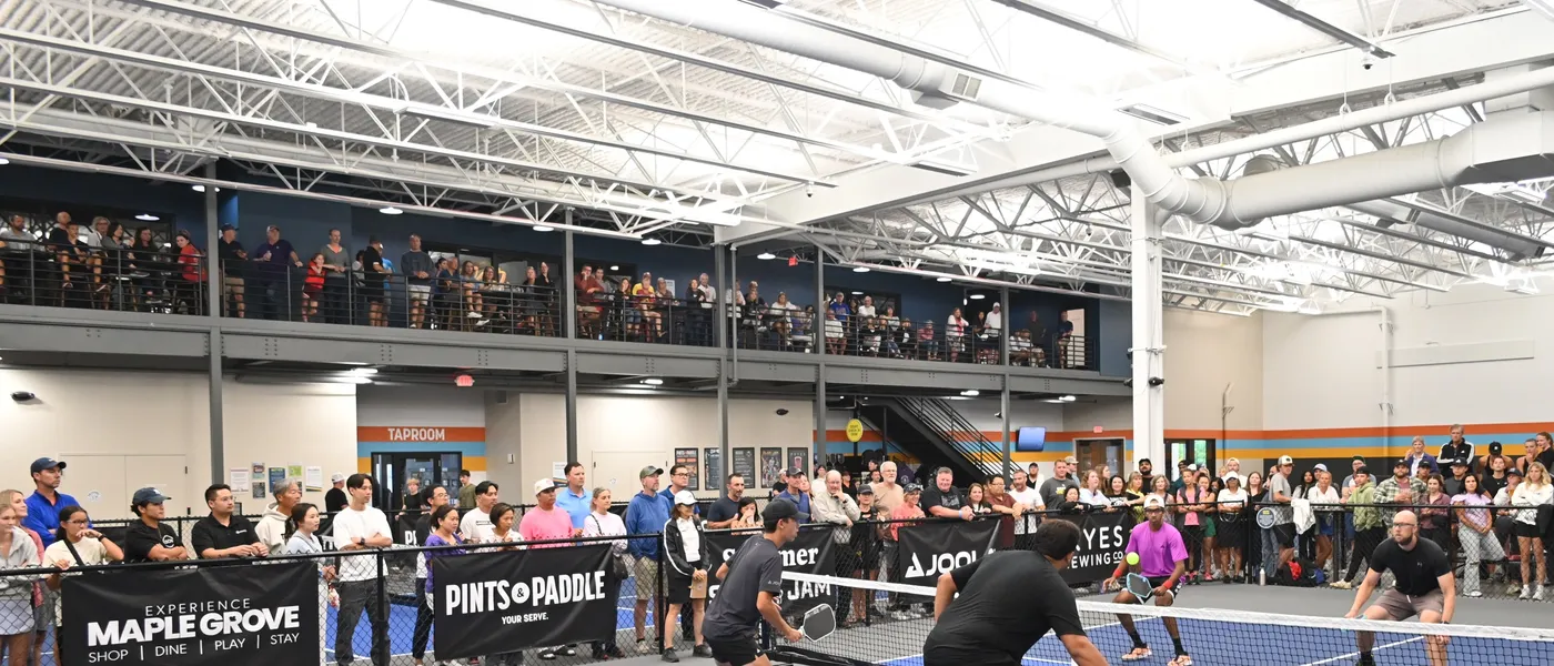 Four pros in a doubles rally at the net during Slam & Jam, spectators two deep along the fence and the mezzanine rail packed end to end above the TAPROOM sign
