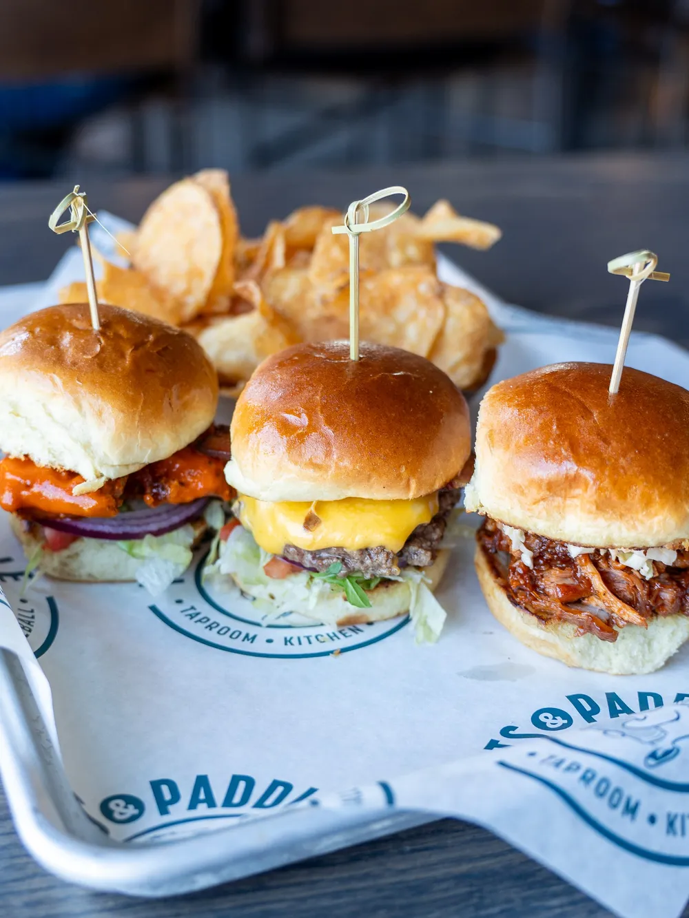 Three sliders on brioche buns—buffalo chicken, a cheeseburger and pulled pork—with kettle chips on a Pints & Paddle tray.