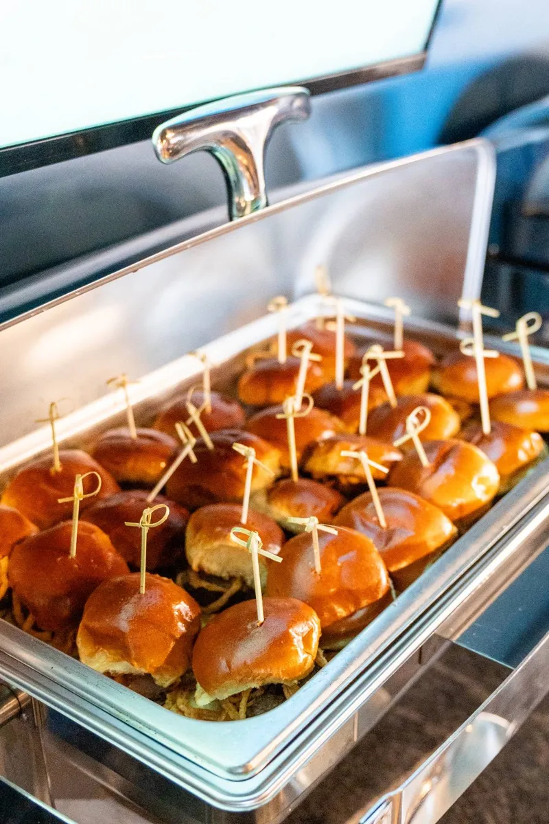 A chafer of brioche sliders on shredded barbacoa, each with a bamboo pick, the lid rolled back