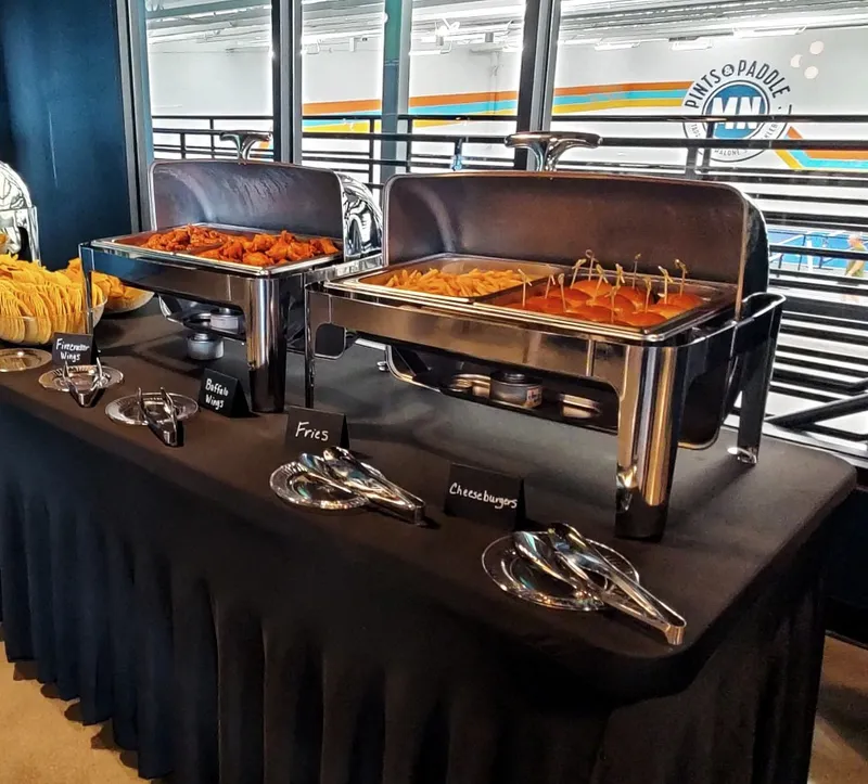 A skirted buffet in the south mezzanine with open chafers of wings, fries and cheeseburger sliders and handwritten name cards, the court windows and MN logo behind