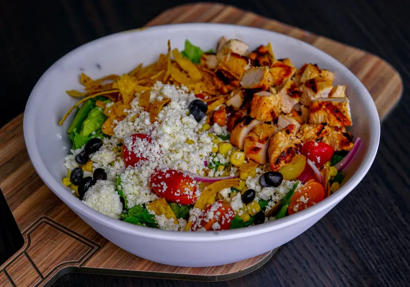 A Southwest chicken salad in a white bowl on a paddle-shaped board: grilled chicken, cotija, black beans, corn, cherry tomatoes and tortilla strips over greens