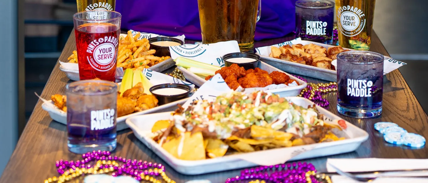 A game day table: nachos, buffalo wings, fries and chicken tenders on branded trays, a pint, a Grape Ape and purple-and-gold beads