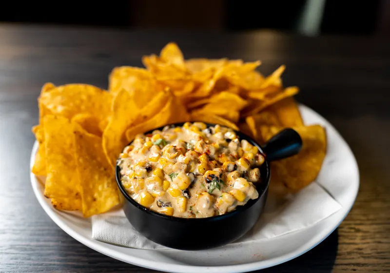 A black skillet of creamy street corn dip dusted with chili powder and cilantro, ringed by tortilla chips on a white plate