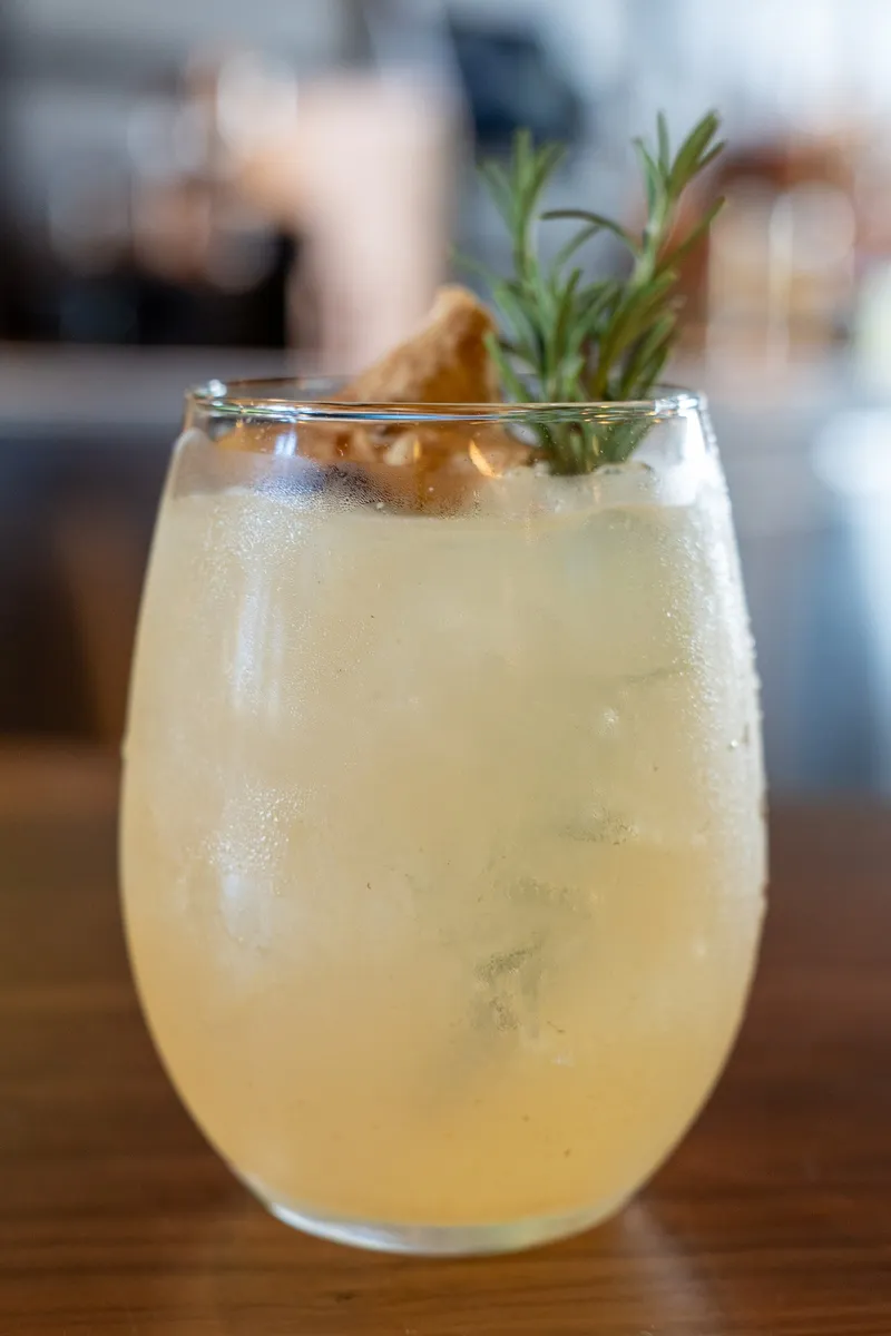 A pale golden Sweater Weather Spritz over ice in a stemless glass with rosemary, a cinnamon stick and dried fruit.