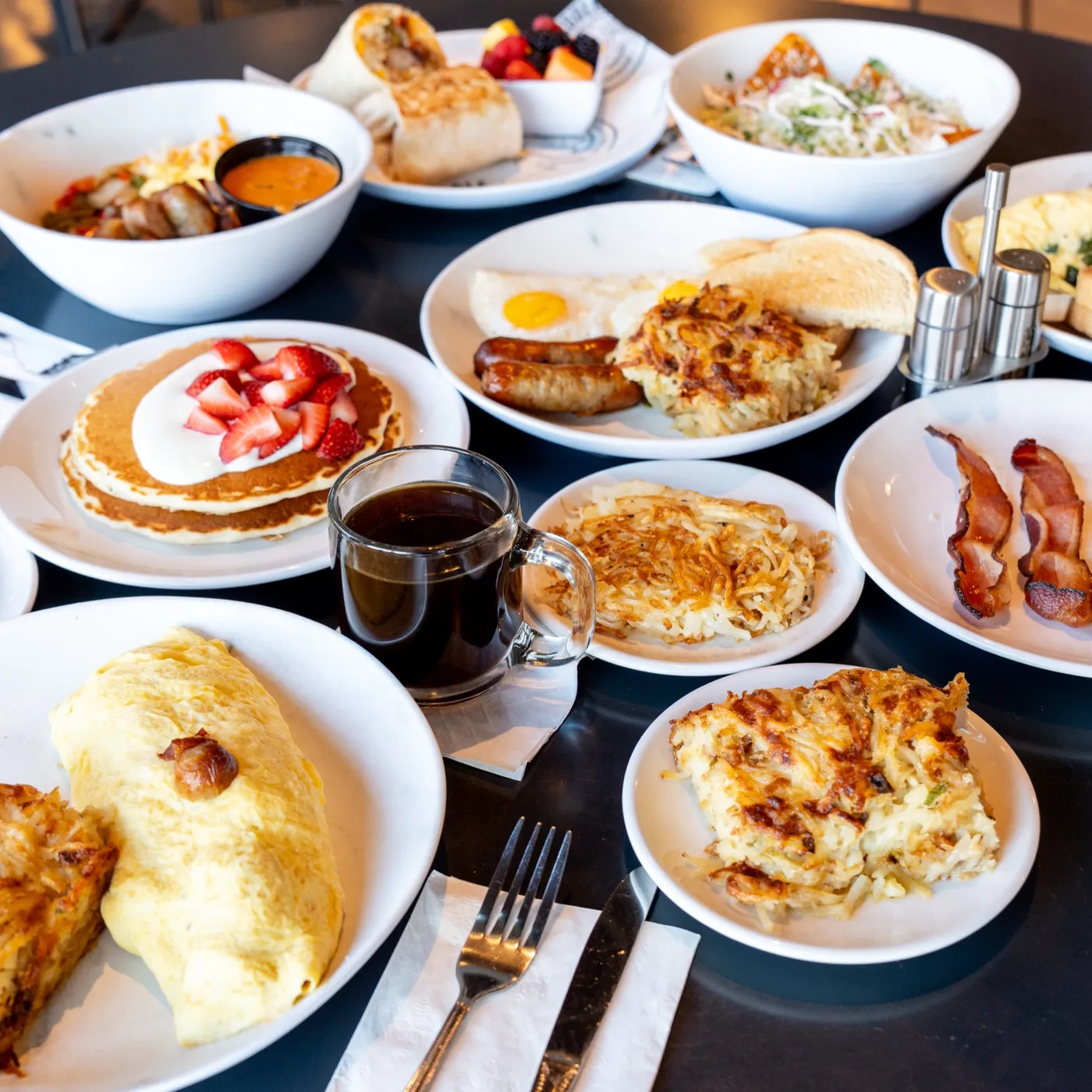 A side view of the breakfast table: a sausage omelette with cheesy hash browns, bacon, pancakes with strawberries, a glass mug of coffee, eggs with sausage and chilaquiles